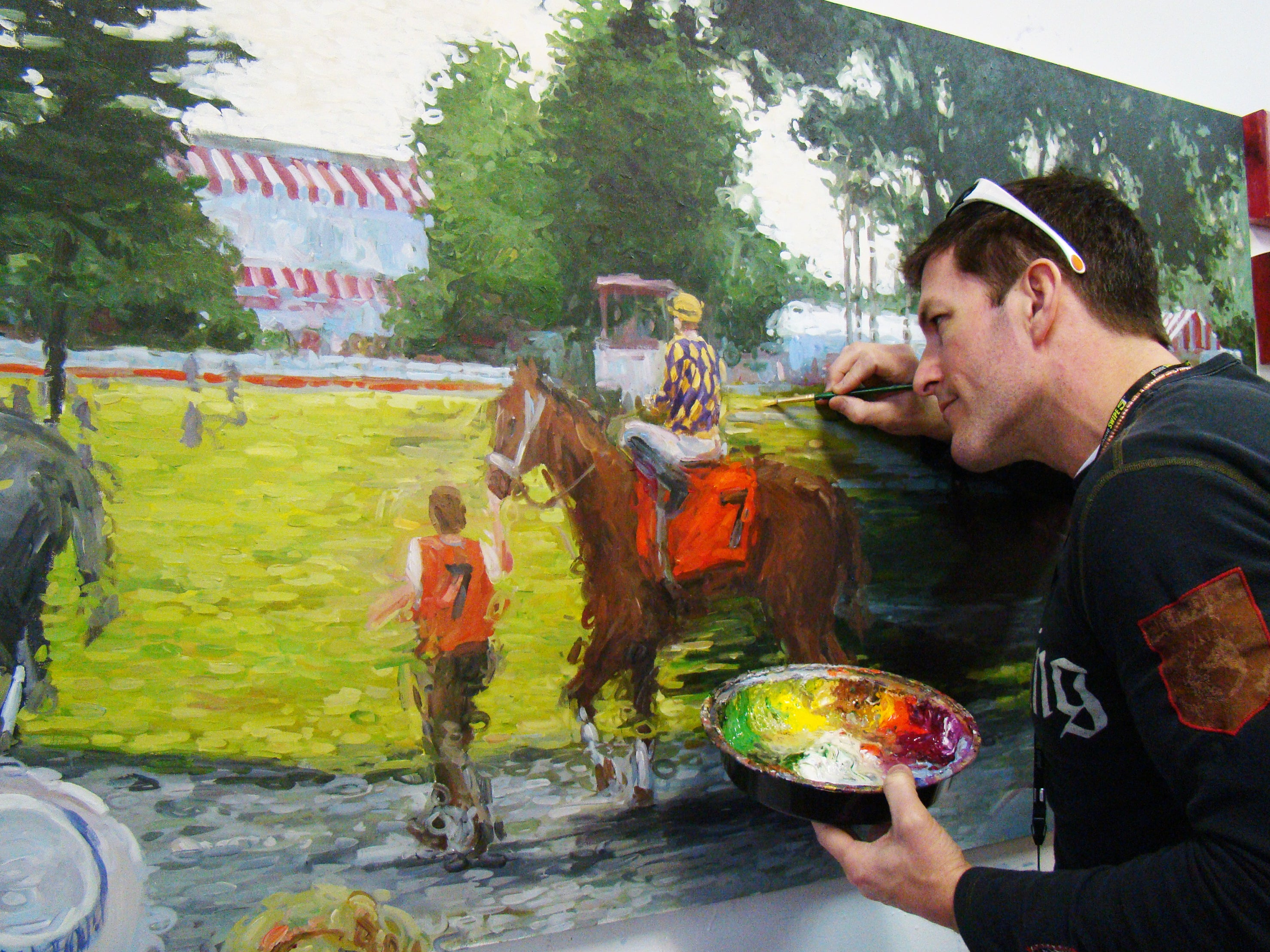 Artist Tom Myott working in his studio on a painting of a scene at Saratoga Race Course. The image shows Myott in profile, meticulously applying paint to a canvas that depicts a jockey on horseback with a stable hand leading them. The painting style is expressive and colorful, with broad brushstrokes capturing the lively atmosphere of the racetrack. In the foreground, Myott holds a palette rich with vibrant hues, emphasizing the artistic process.