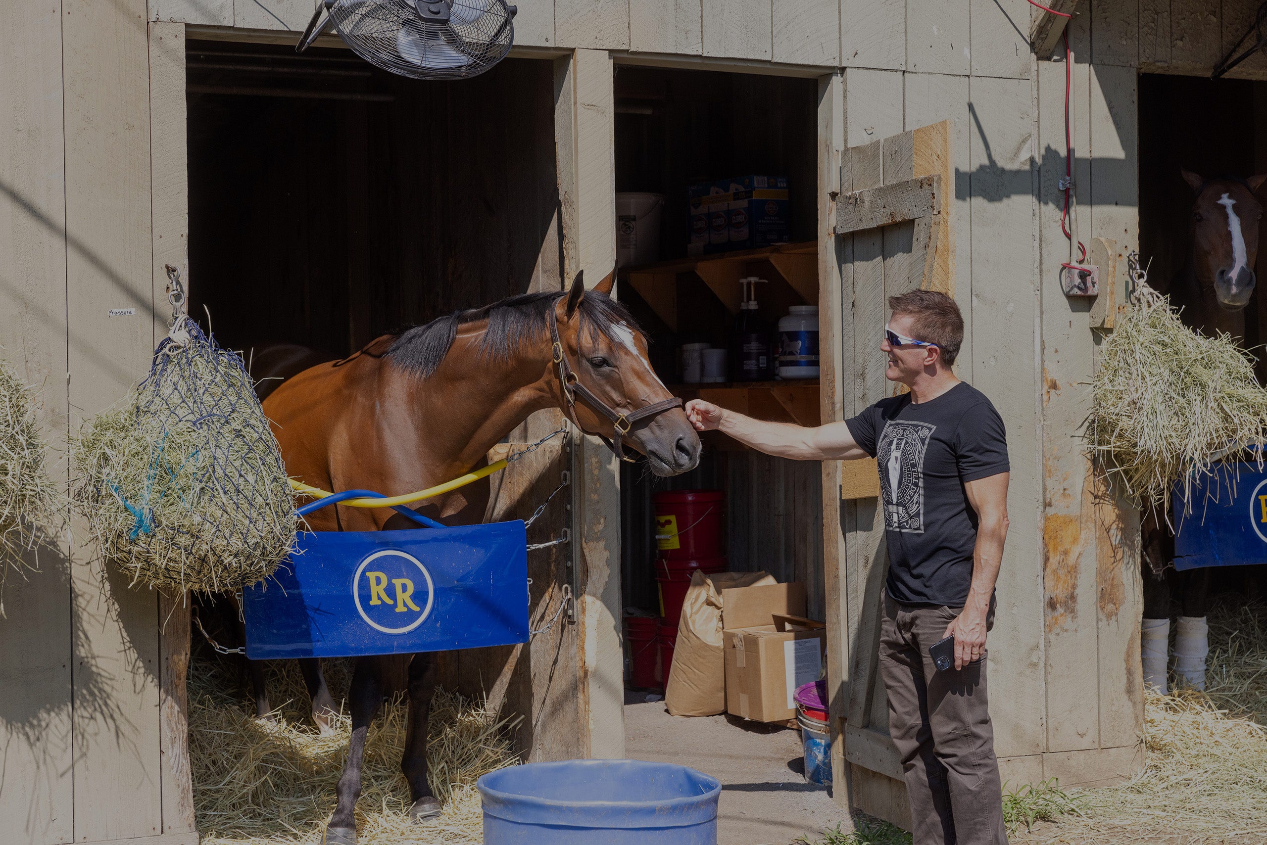 Artist Tom Myott standing beside a stable, gently petting a racehorse. The horse, with a sleek bay coat, is partly inside the stall, which is equipped with hay and other horse care supplies. Myott, appears relaxed and engaged with the horse, capturing a moment of connection between the animal and the artist known for his equine paintings.