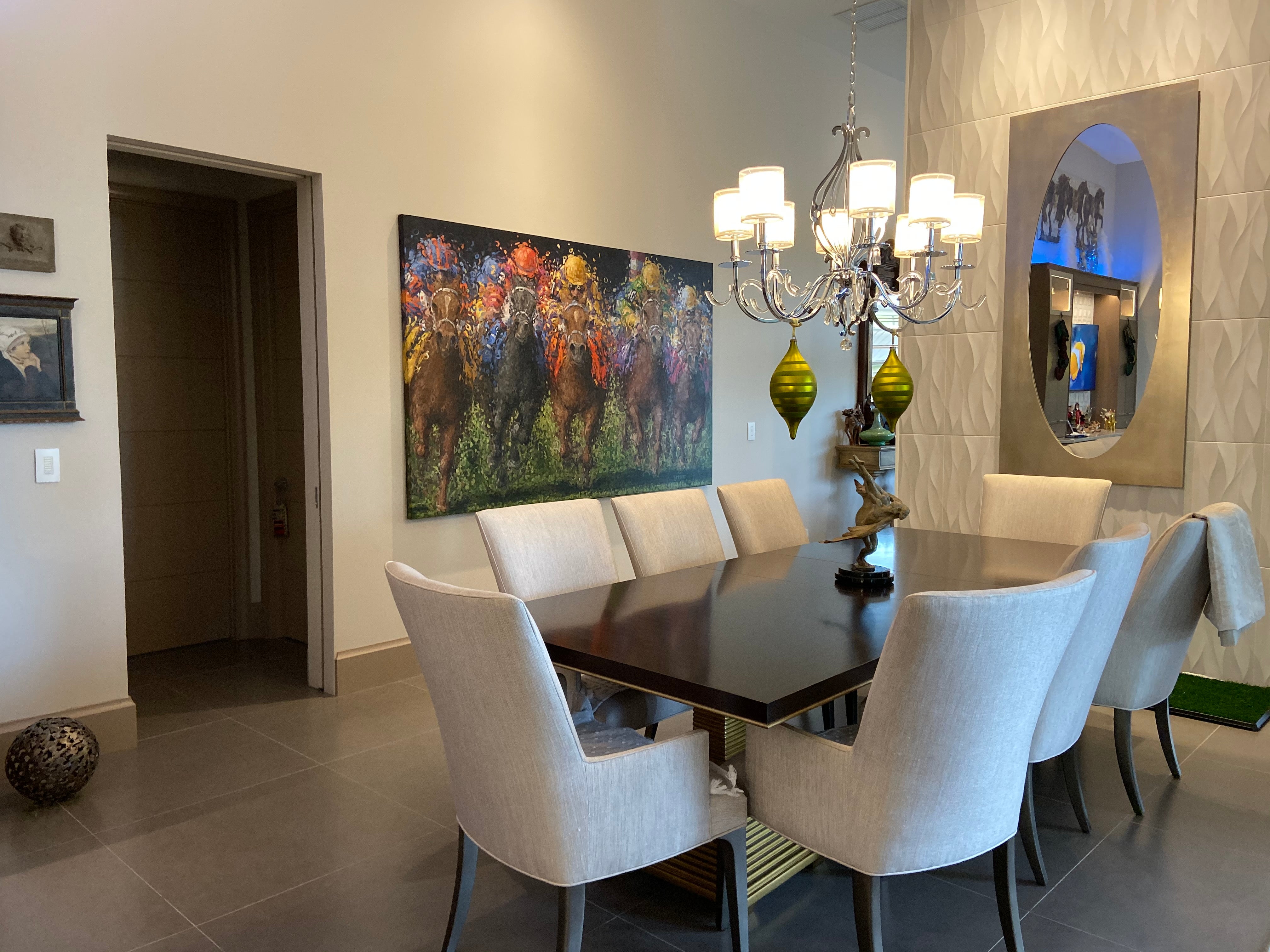 A contemporary dining room featuring a large, colorful painting by Tom Myott on the wall, depicting a vibrant horse race. The artwork's lively brushstrokes and rich colors complement the room's modern aesthetics, which include a sleek dark wood dining table surrounded by light fabric chairs. An elegant chandelier hangs above the table, and a decorative mirror on the opposite wall reflects the room's natural light. A sculpture centerpiece and a textured wall panel add to the sophistication of the space.