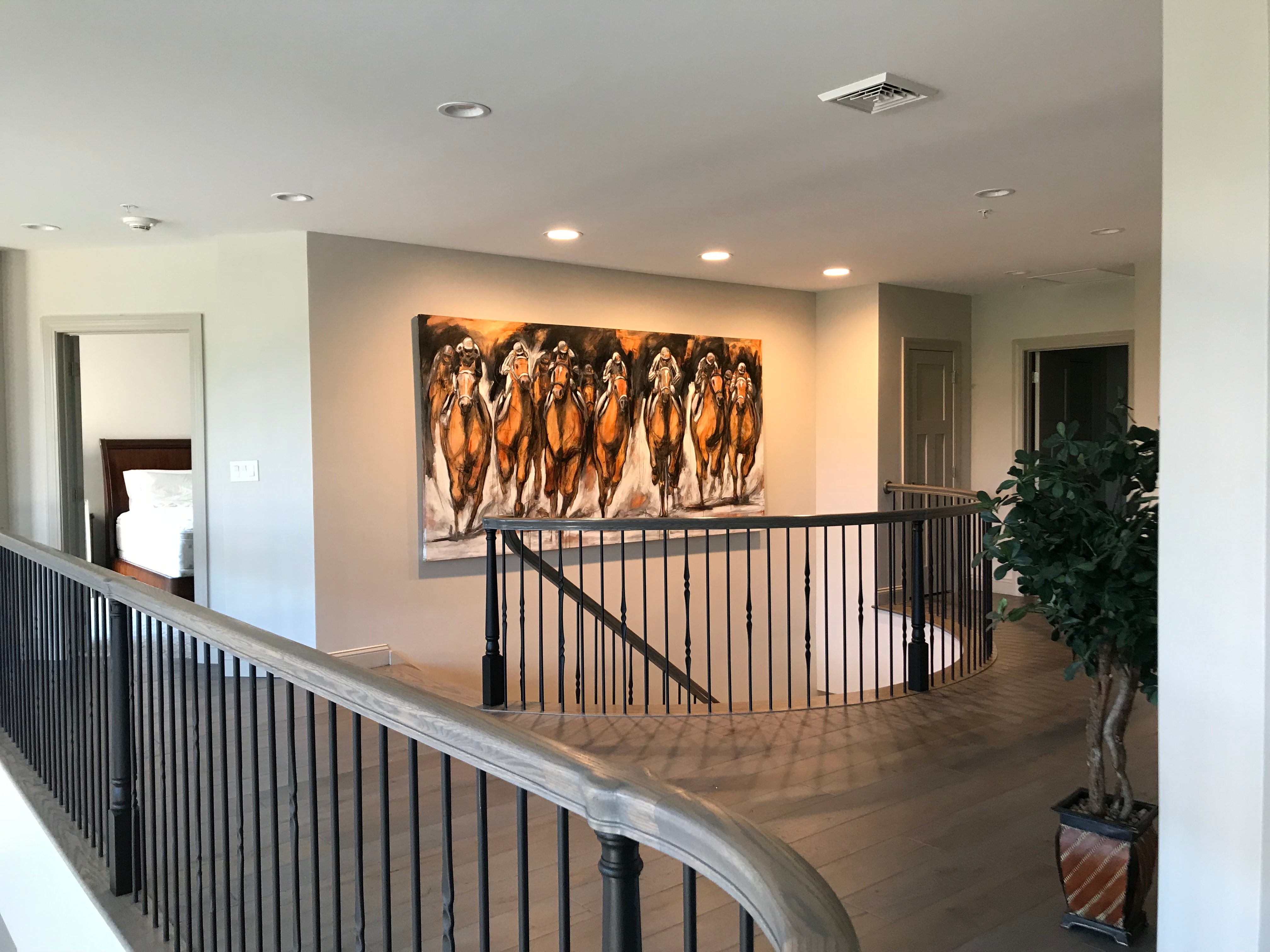 A spacious residential interior with a large painting by Tom Myott hanging prominently on a wall. The painting features a group of powerful, spirited horses captured in warm earth tones, showcasing Myott's characteristic bold brushstrokes. The artwork is a focal point above the staircase landing, flanked by two doors and complemented by a potted plant to the right, giving the modern home a touch of equestrian elegance.