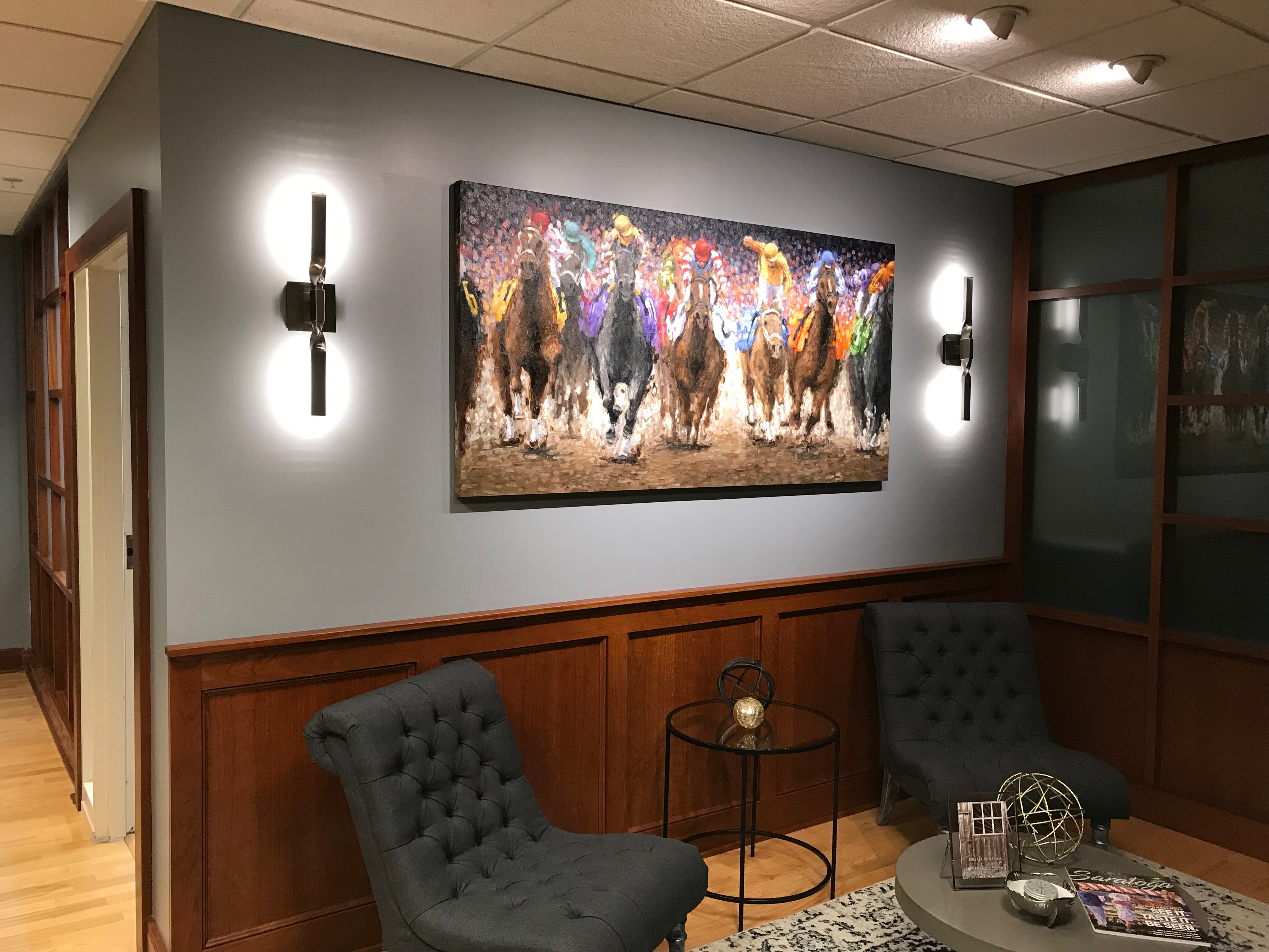 In a client's office, a striking painting by Tom Myott is displayed on a light grey wall above a wood-paneled wainscoting. The painting, illuminated by two modern wall sconces, depicts a lively scene of colorful horses in motion, characteristic of Myott's style. The office is furnished with two dark upholstered chairs and a round table with decorative objects, creating a sophisticated and inviting area for contemplation or conversation, with the artwork serving as a compelling centerpiece.
