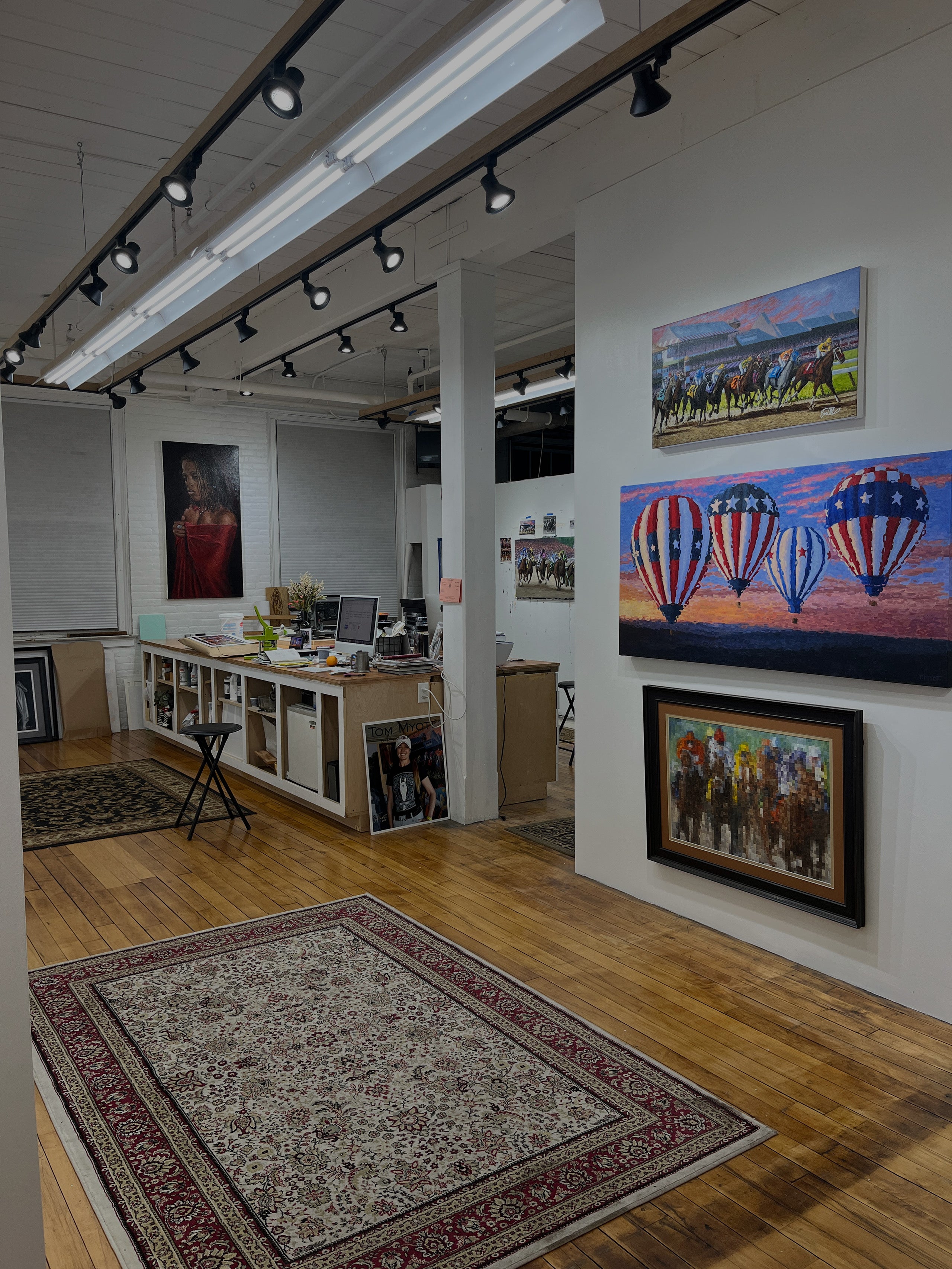 Interior view of Tom Myott's gallery and studio space located in The Shirt Factory in Glens Falls, NY. The studio is well-lit with track lighting and features a polished hardwood floor with a large ornate area rug. Various artworks are displayed on the white walls, including a prominent painting of hot air balloons with American flag motifs. The workspace contains a desk with a computer, art supplies, and canvases, creating a creative and productive atmosphere.