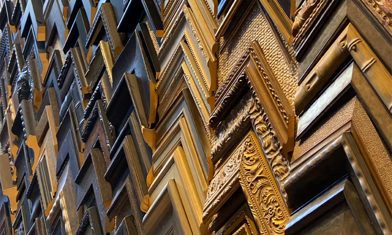 A close-up photo showcasing a diverse array of picture frames offered by Tom Myott. The collection includes a variety of styles and finishes, from sleek modern black and silver frames to ornate, classical designs with intricate gold leaf detailing. The multitude of choices highlights the custom framing options available for artwork, each frame capable of complementing a different artistic style or home decor.