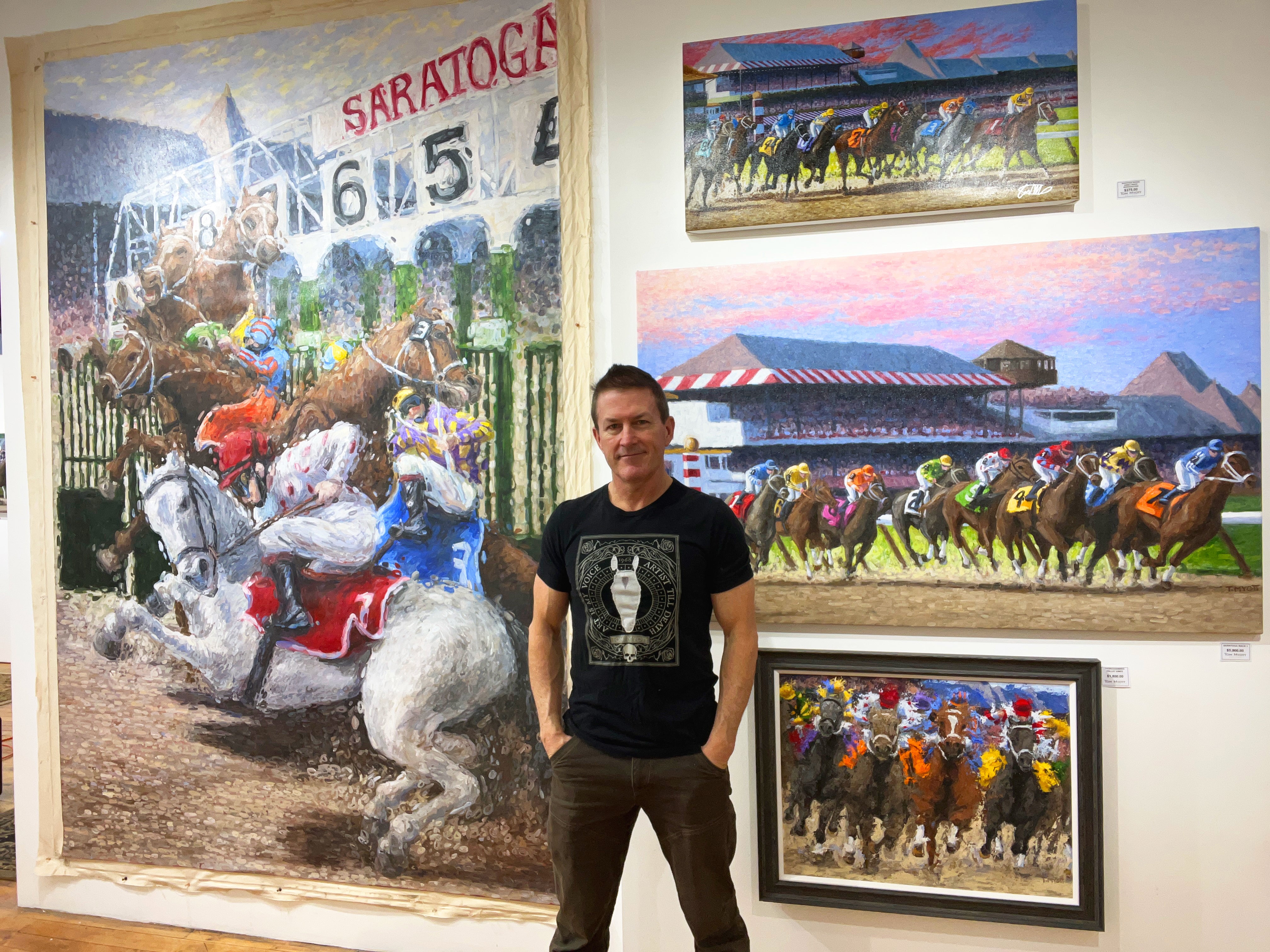 Artist Tom Myott poses in his studio with a proud smile, standing in front of his vibrant paintings of horse racing scenes at Saratoga Race Course. The paintings capture dynamic moments of races, featuring jockeys in colorful silks on galloping horses, with one large painting prominently displaying the iconic Saratoga starting gate. The artwork exhibits Myott’s expressive brushwork and vivid use of color, showcasing his talent in equine art.