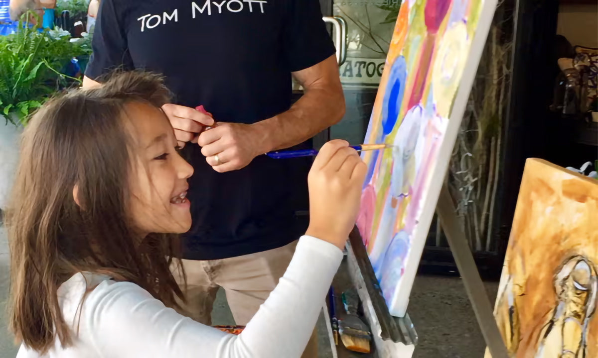 Tom Myott engages in a community art event, painting outdoors on a sidewalk in Saratoga, sharing a canvas with a young child who is also painting. The child, in a white long-sleeve shirt, smiles brightly as she adds brushstrokes to the colorful abstract work. Myott, seen from behind, appears focused on guiding the child's hand, fostering a collaborative artistic experience.