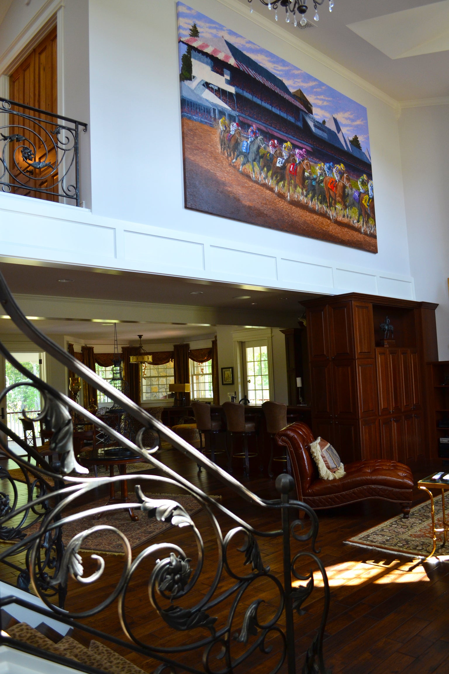 An elegant home interior with a grand Tom Myott painting mounted high on the wall above the entrance to a traditional dining room. The painting vividly portrays a horse racing scene at Saratoga, filled with the energy of mid-race action. Below, the home features rich wood paneling, a leather sofa, and an ornate wrought-iron staircase railing, which adds to the luxury of the space. The artwork serves as a commanding focal point, visible from both the lower and upper levels of the home.