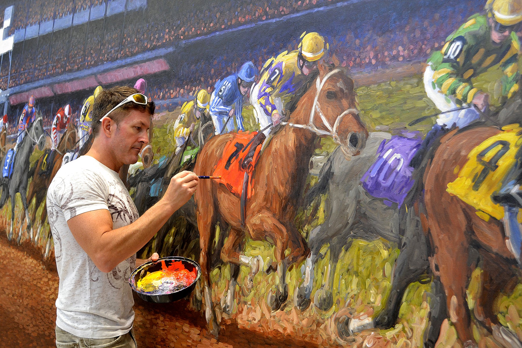 Artist Tom Myott is painting a large-scale commission piece. He is in the process of detailing a horse racing scene, with a focus on a brown horse with the number seven. Myott, in a casual white shirt and holding a palette full of vibrant colors, is painting with concentration and precision. The artwork captures the dynamic energy of the race, with jockeys in colorful silks riding their mounts, depicted in Myott’s signature expressive style.